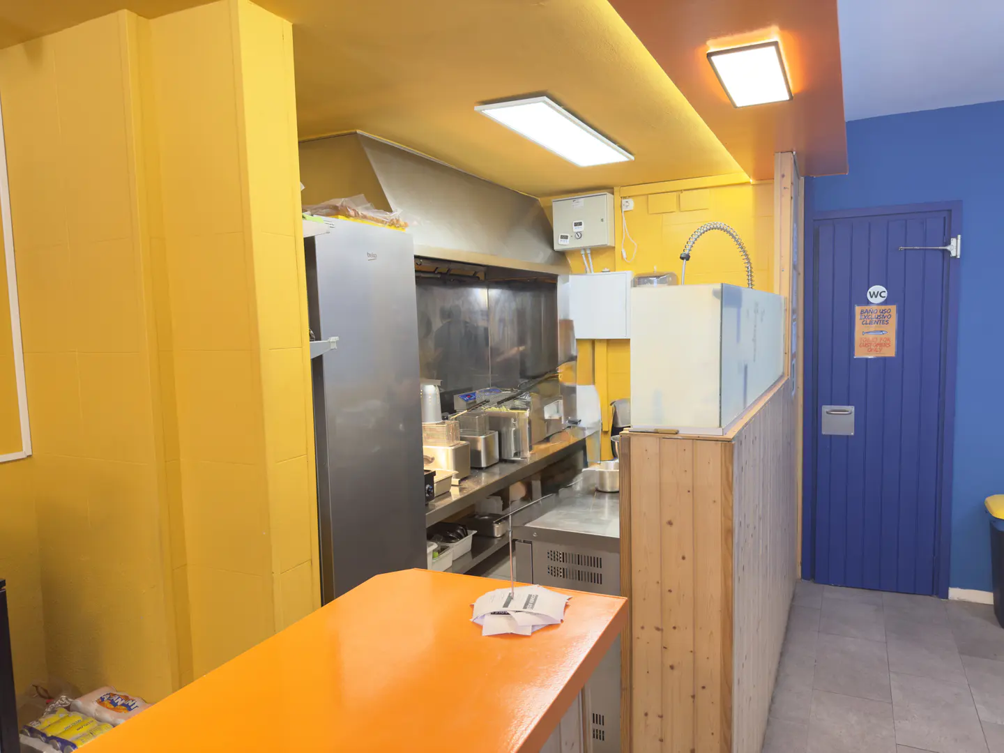 A commercial kitchen with yellow walls, stainless steel appliances, and an orange counter. A blue door with a WC sign is visible.