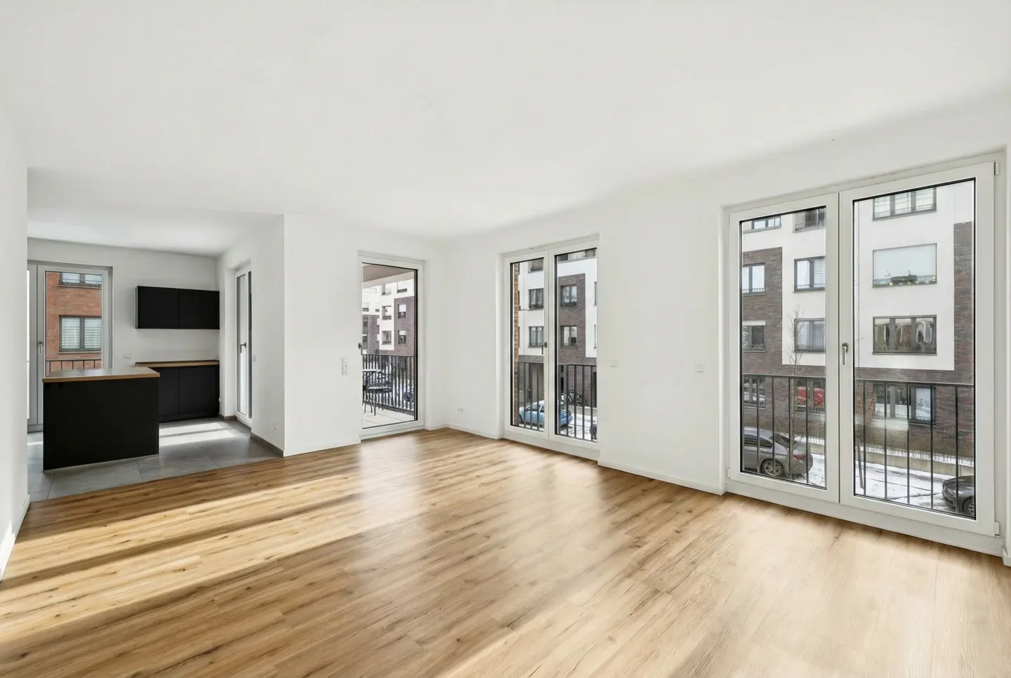 Bright, modern apartment with wood floors, white walls, and large windows. A black kitchen island is visible in the background.