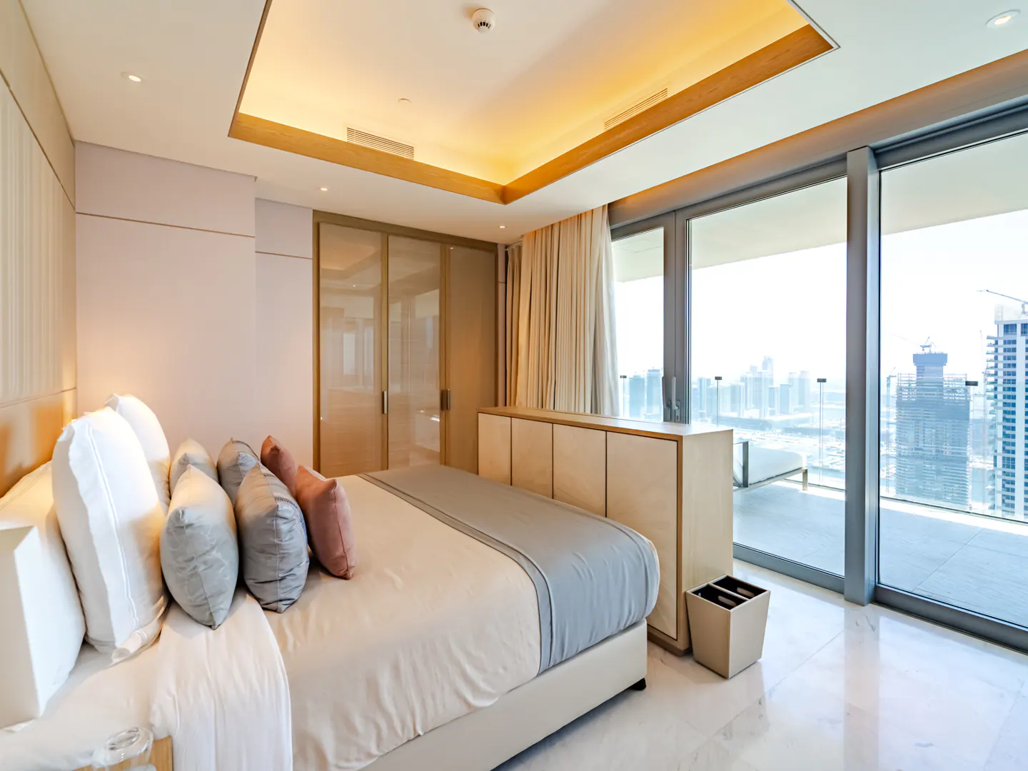 Modern bedroom with a plush bed, neutral decor, and floor-to-ceiling windows offering a panoramic city skyline view.