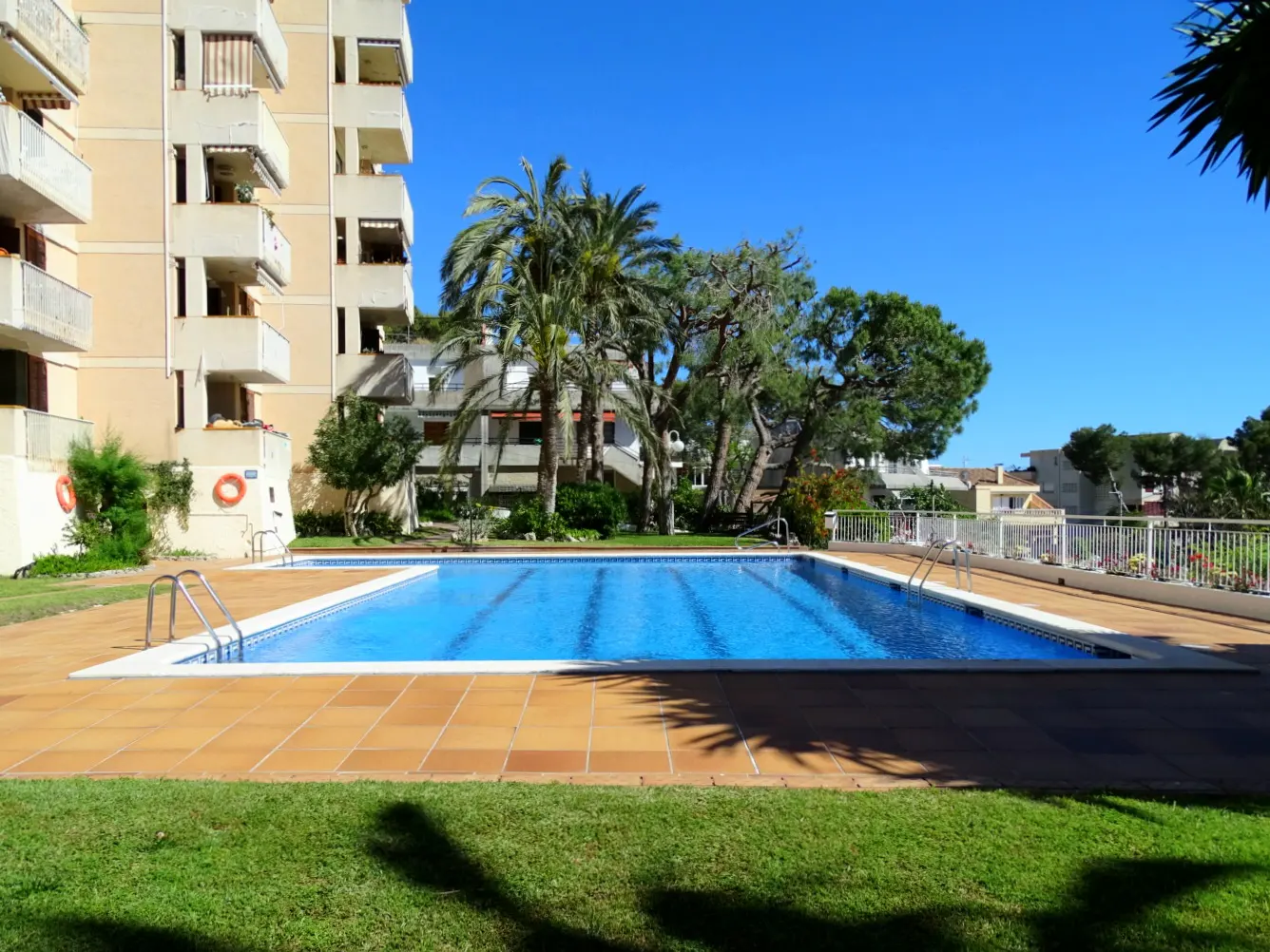 Outdoor pool with blue water and brown tile surround, next to a light yellow apartment building and green lawn under a clear blue sky.