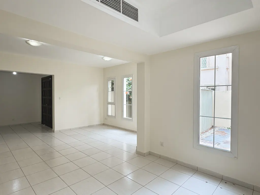 Bright, empty room with white walls, tile floor, and windows. A dark door is visible in the background.