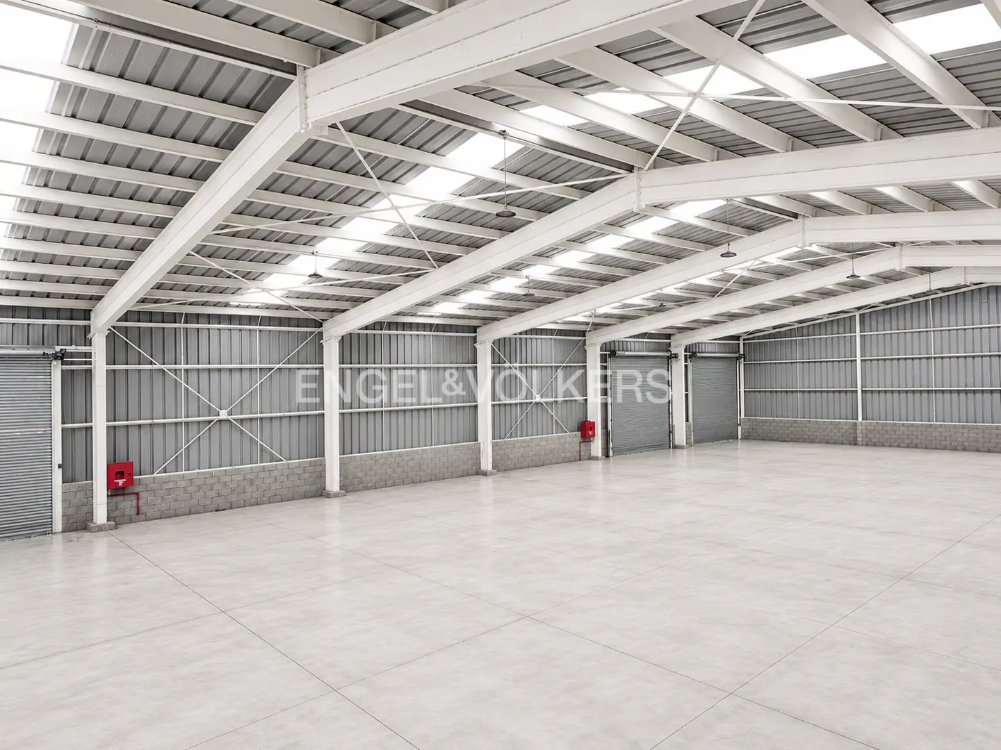 Empty, spacious industrial warehouse with high ceilings, white steel beams, concrete floors, and grey metal walls.