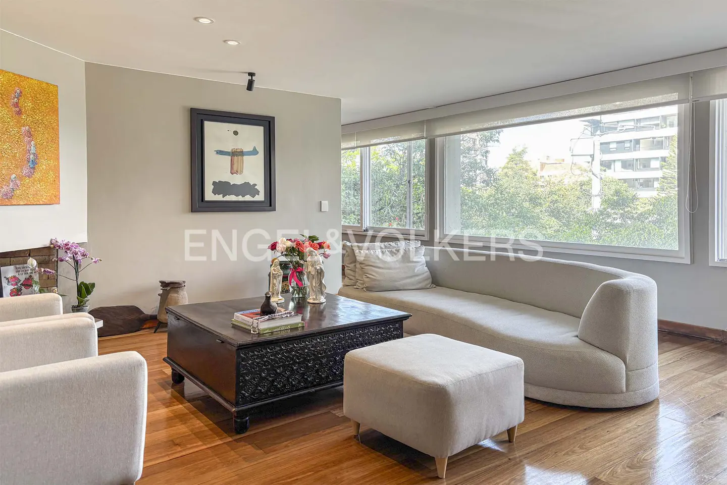 Living room with hardwood floors, a curved white sofa, and a dark wood carved coffee table. Large windows overlook green trees.
