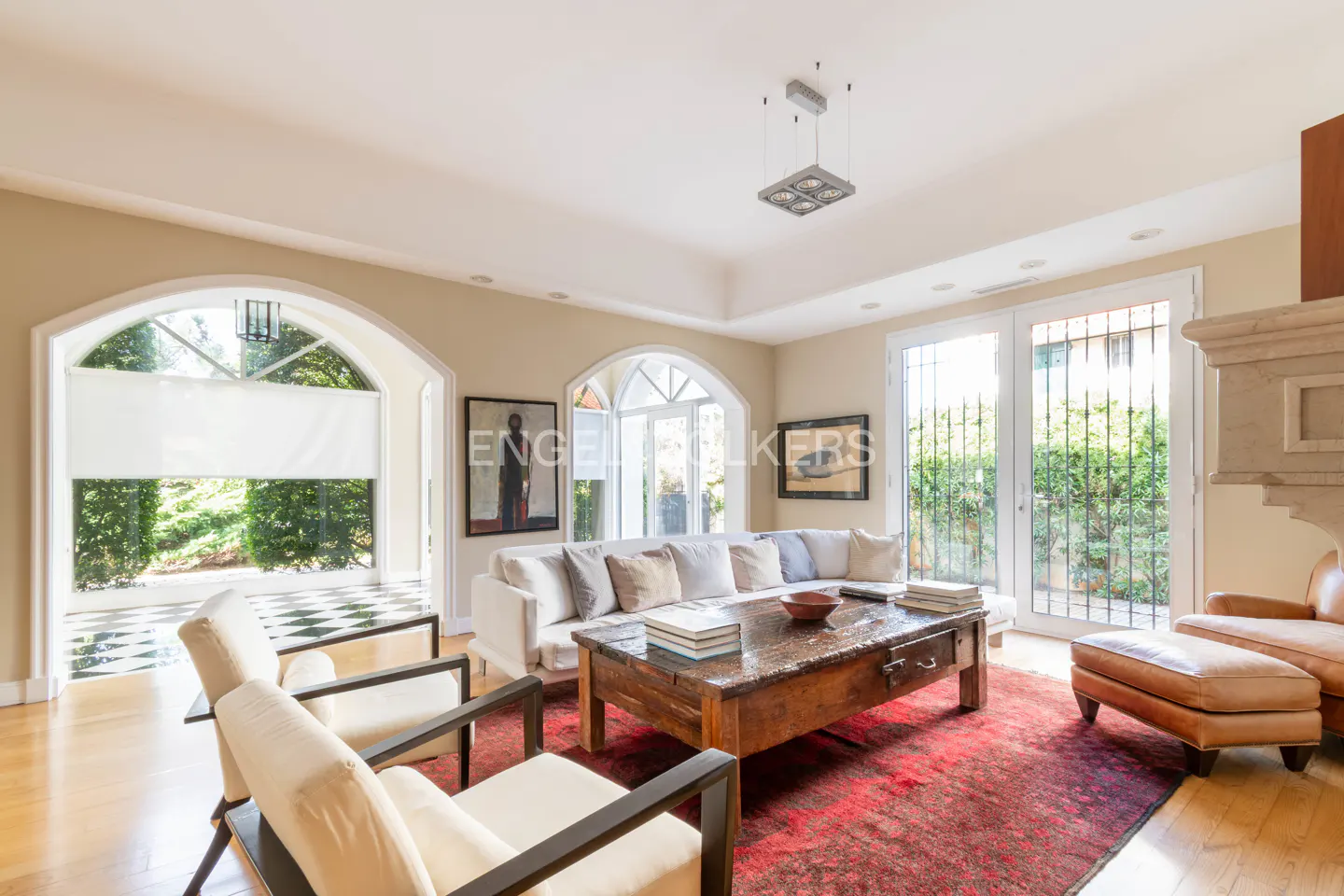 Bright living room with white sofa, wood coffee table, and red rug. Arched windows overlook greenery. Two chairs face the sofa.