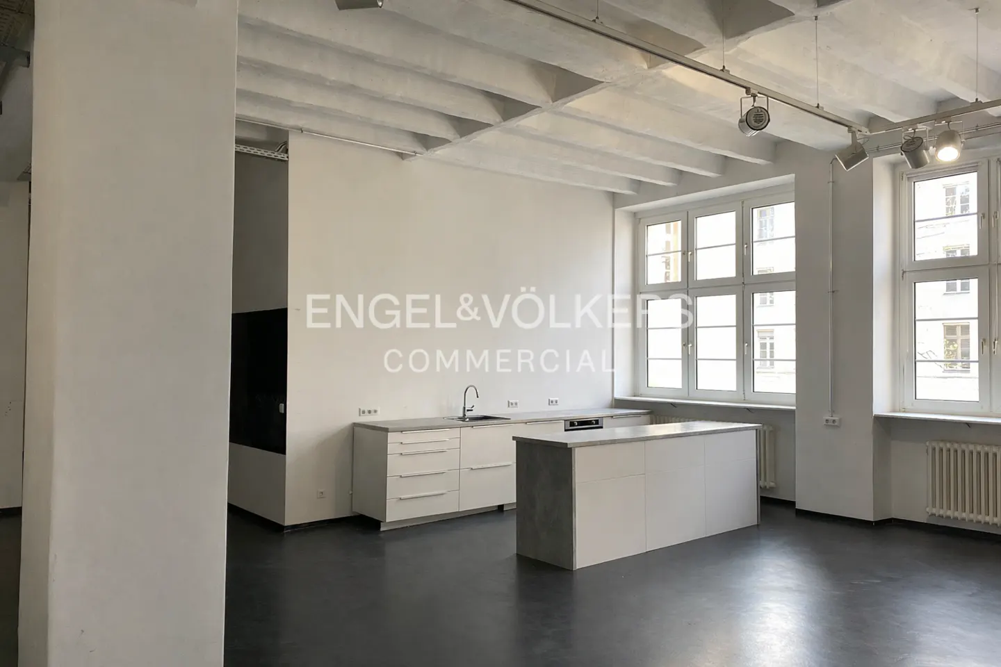 Modern white kitchen in a spacious loft with high ceilings, large windows, and dark flooring.