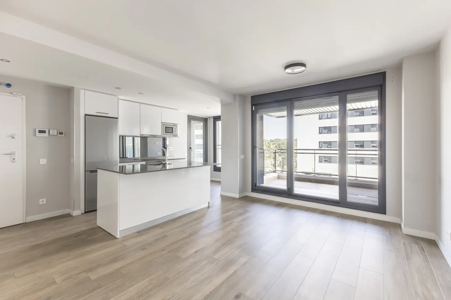 Bright, modern apartment interior with wood floors, white kitchen island, stainless steel appliances, and sliding glass doors to a balcony.