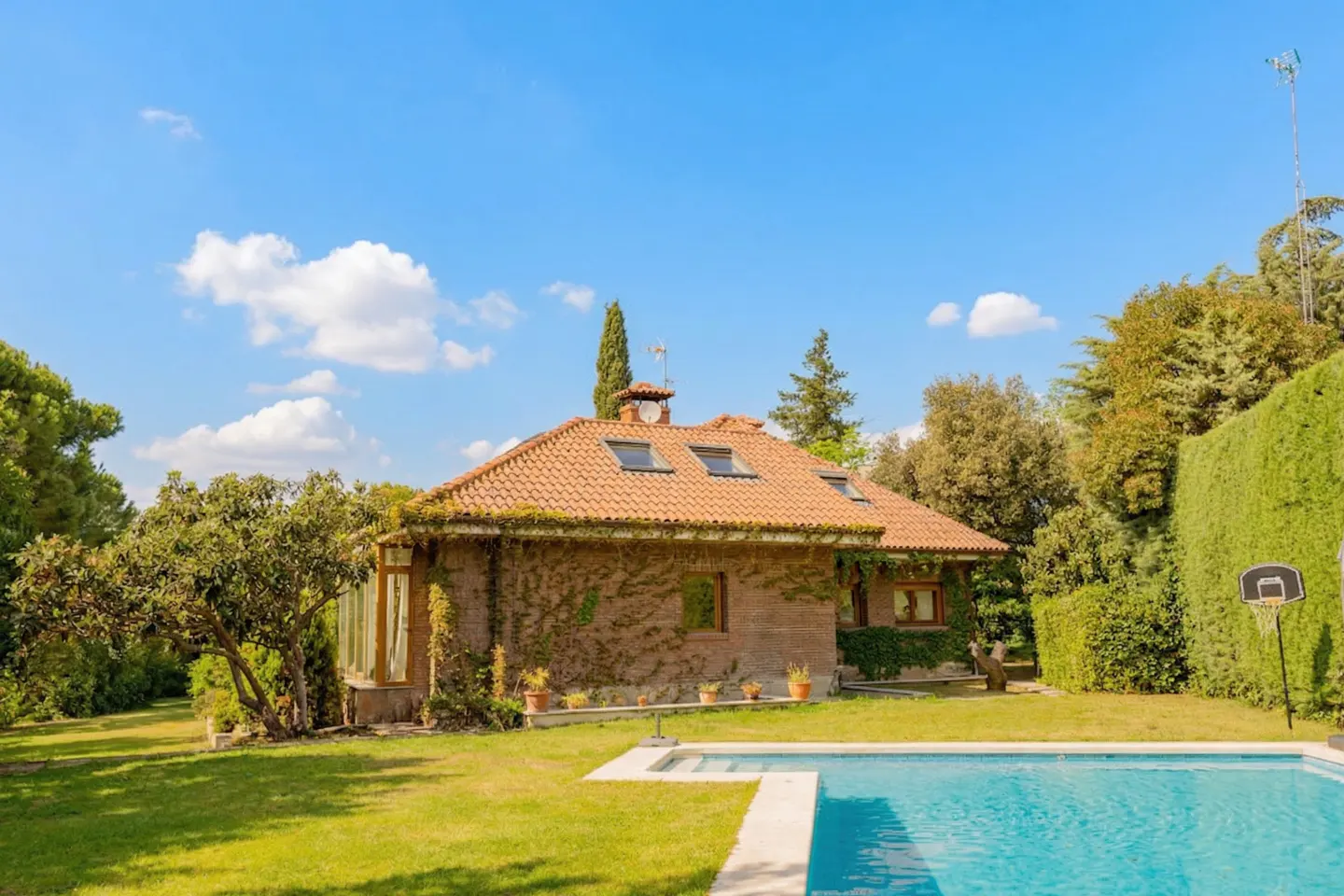 Exterior view of a brick house with a red tile roof, ivy, a pool, and a green lawn under a blue sky.