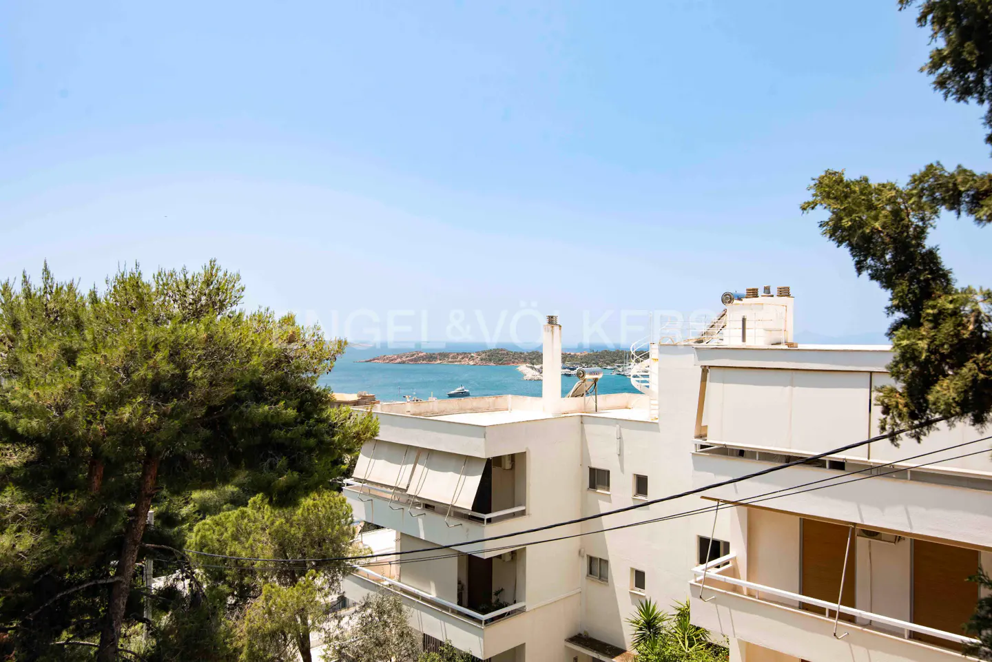 View of a white building with balconies overlooking the sea and islands under a blue sky, framed by green trees.