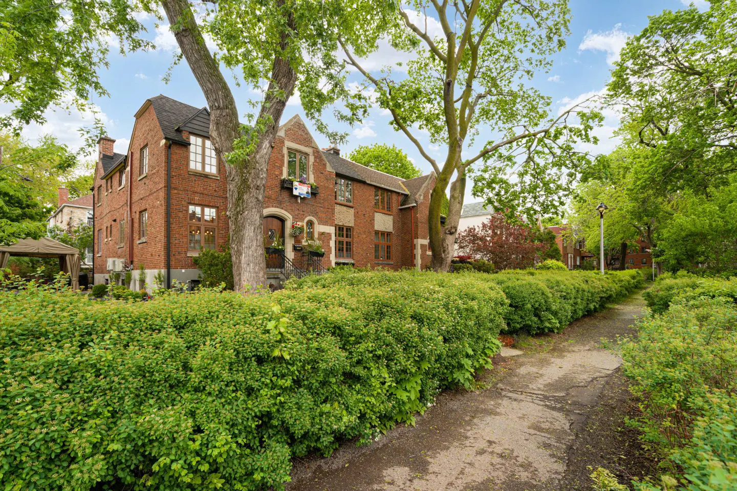A two-story red brick house with a black roof, surrounded by green trees and bushes. A stone path leads to the house.