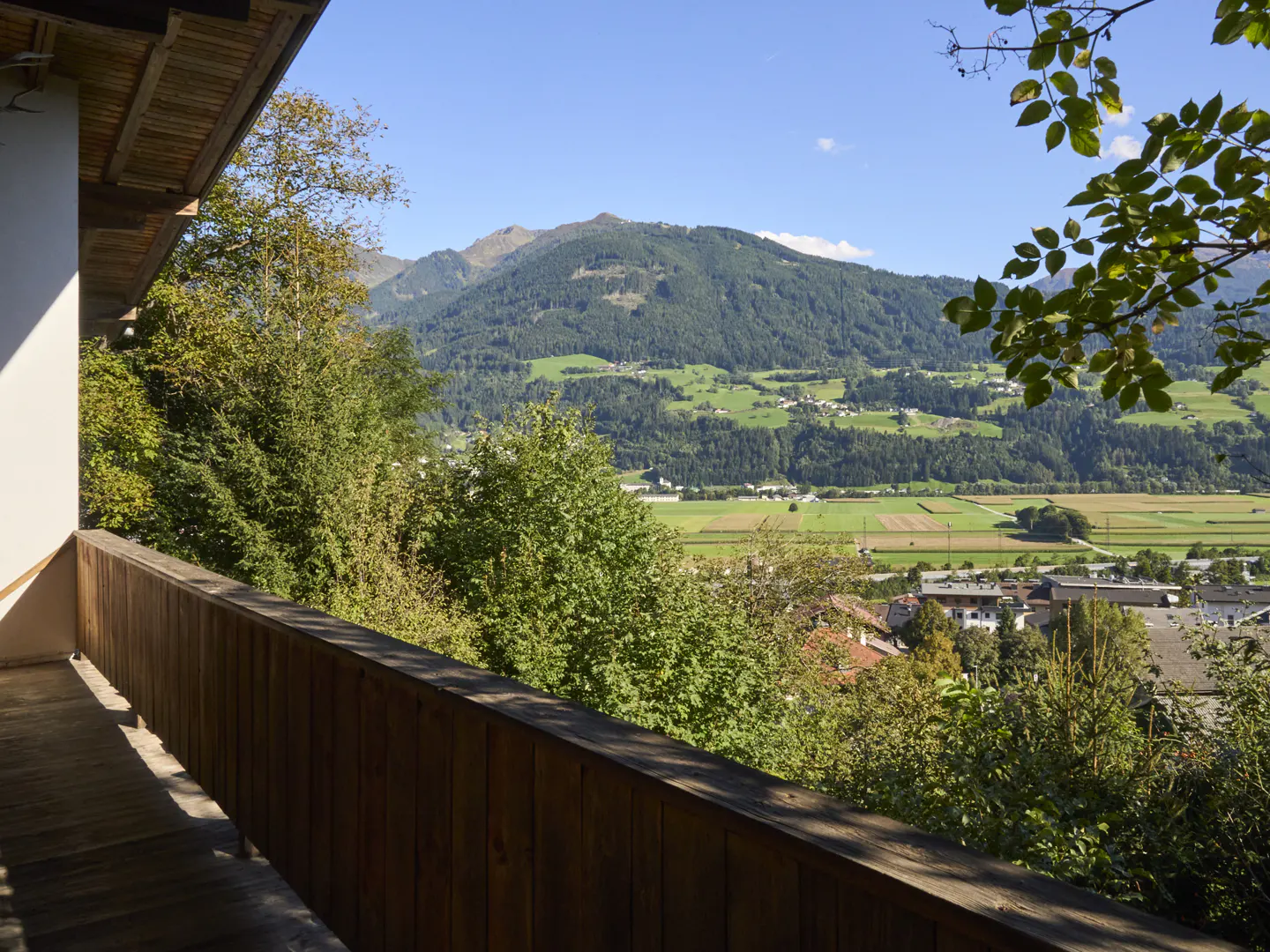 View from a wooden balcony overlooking green trees, fields, and mountains under a blue sky.