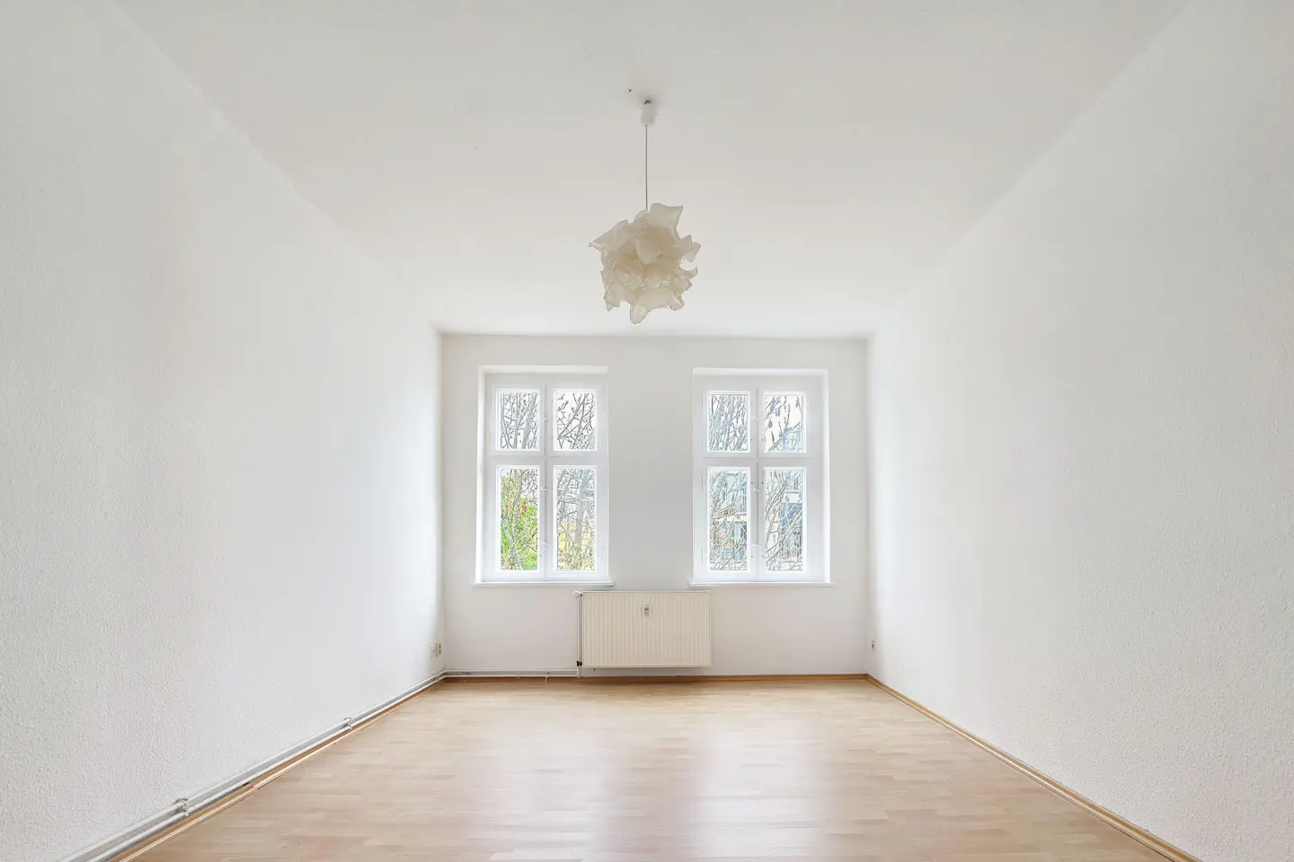 Bright, empty room with hardwood floors, white walls, and two windows. A modern, white pendant light hangs from the ceiling.