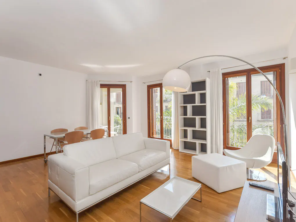 Bright living room with white sofa, chair, and ottoman on a wood floor. Large windows and a modern floor lamp add to the airy feel.