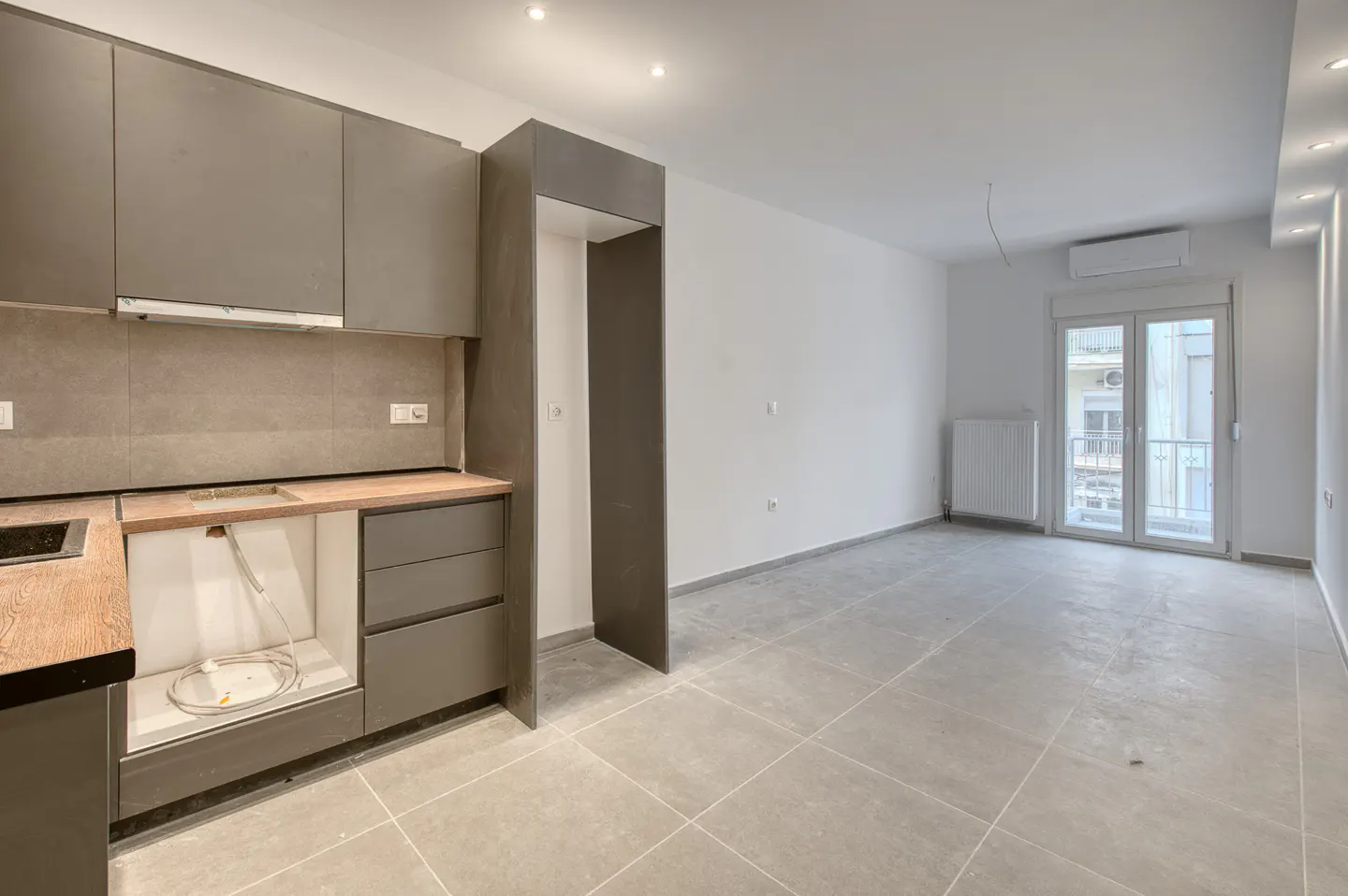 A bright, empty apartment with gray tile floors, white walls, and gray kitchen cabinets. A door leads to a balcony.