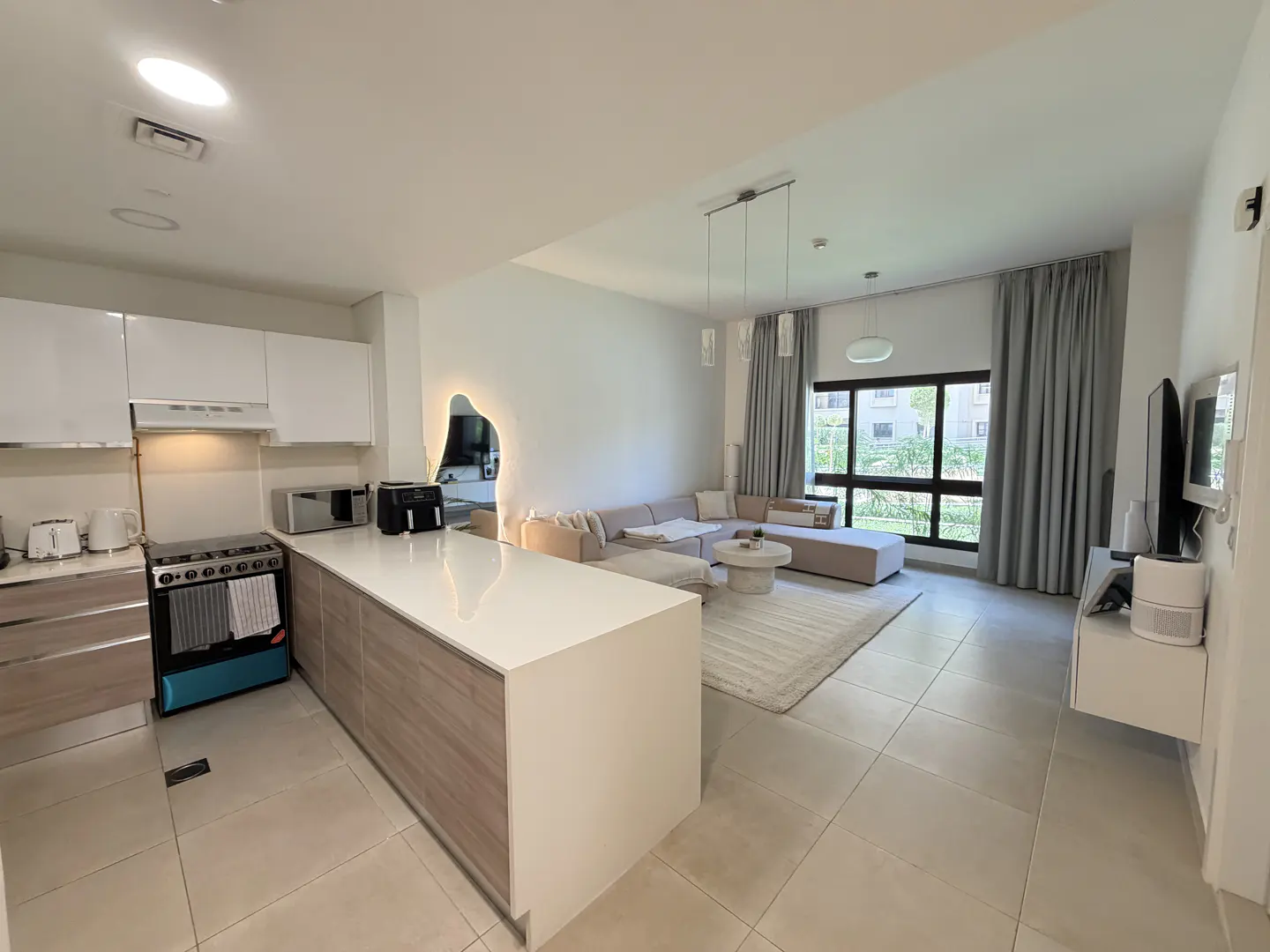 Open-concept living space with white cabinets, island, and beige sectional sofa. Large window with gray curtains.
