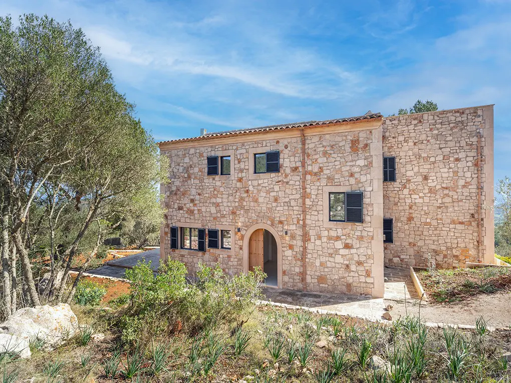 Two-story stone house with black shutters and an arched doorway, set against a blue sky.
