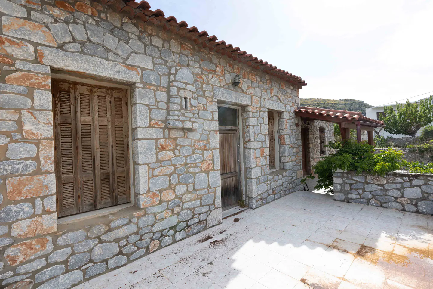 Exterior view of a stone house with a red tile roof, wooden shutters, and a tiled patio.
