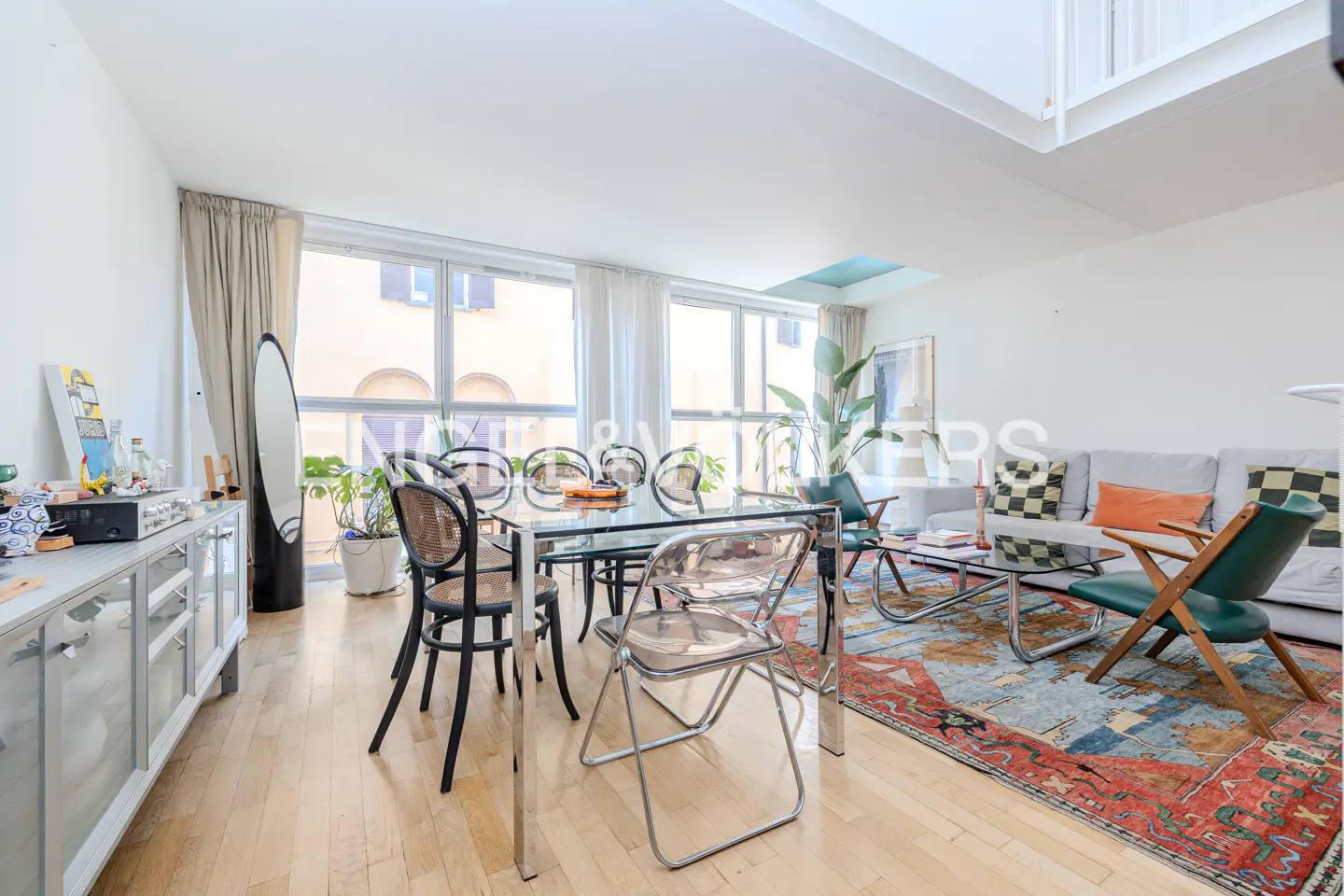 Bright living and dining area with a glass table, modern chairs, a colorful rug, and a sofa under a high ceiling.