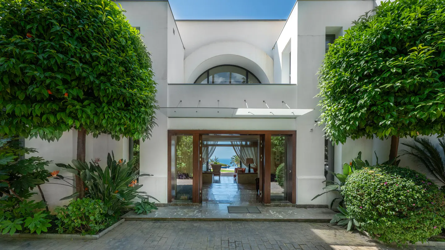 Front view of a white house with an open door, green trees, and a glimpse of the ocean in the background.