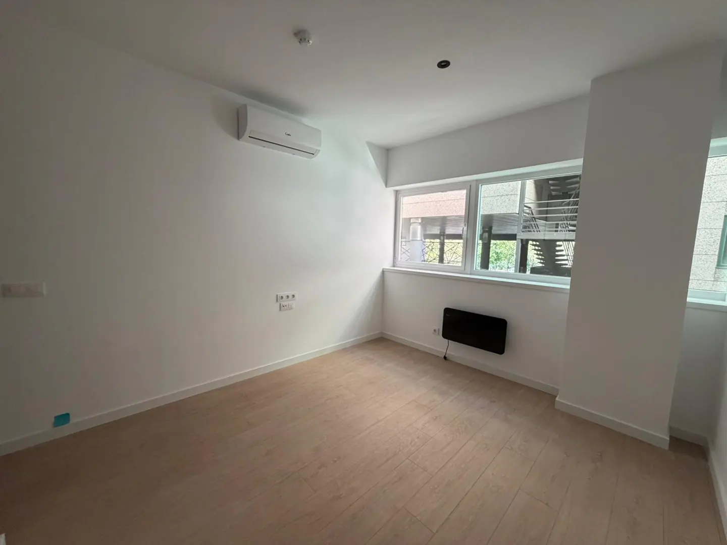 Bright, empty room with light wood floors, white walls, and large windows. An air conditioner is mounted high on the wall.