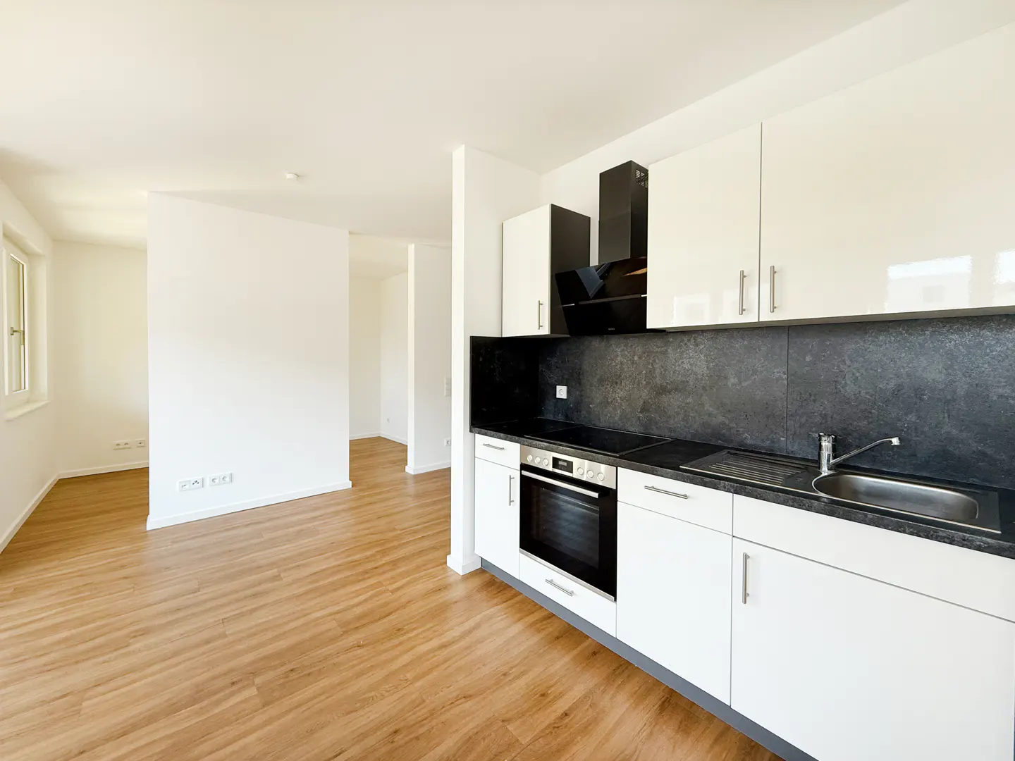 Bright, modern kitchen with white cabinets, black countertops, and wood floors. Oven, sink, and open doorway visible.