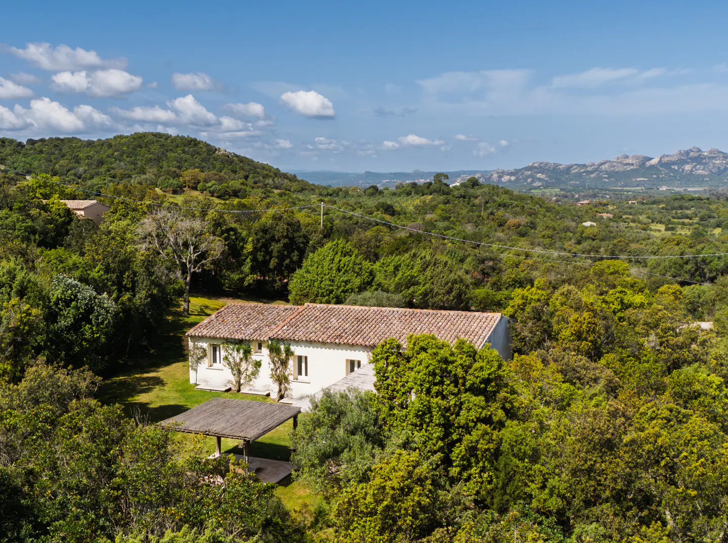 A white house with a red tile roof is surrounded by green trees and hills under a blue sky.