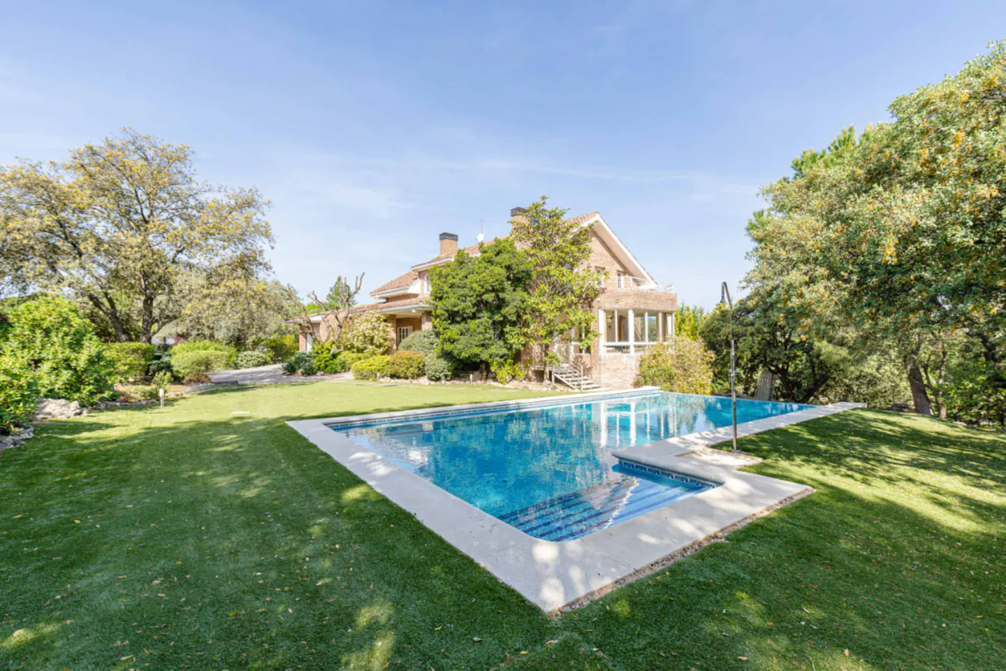 A large, rectangular swimming pool with blue water sits in a green lawn in front of a two-story brick house.