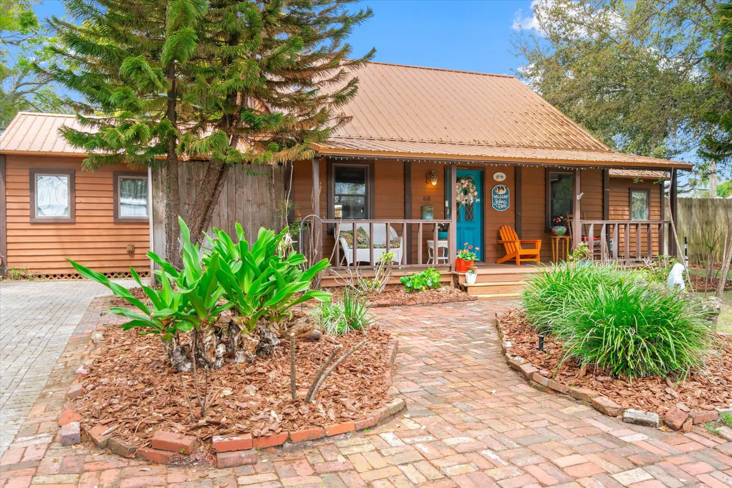 A brown house with a metal roof, a blue door, and a brick walkway. The porch has white chairs and an orange Adirondack chair.