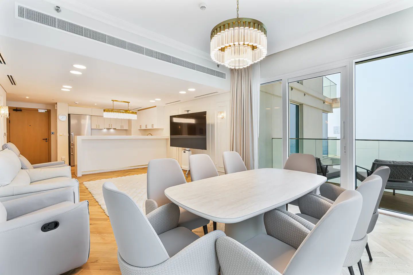 Bright, modern apartment interior with a dining table, chairs, and a view of the balcony. A chandelier hangs above the table.