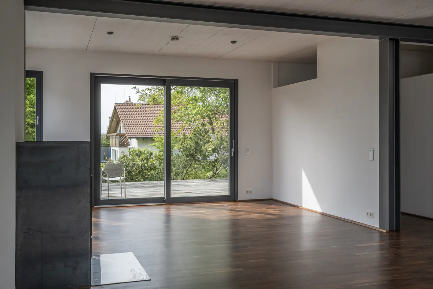 An empty room with dark wood floors, white walls, and a sliding glass door to a deck with a view of a house and trees.
