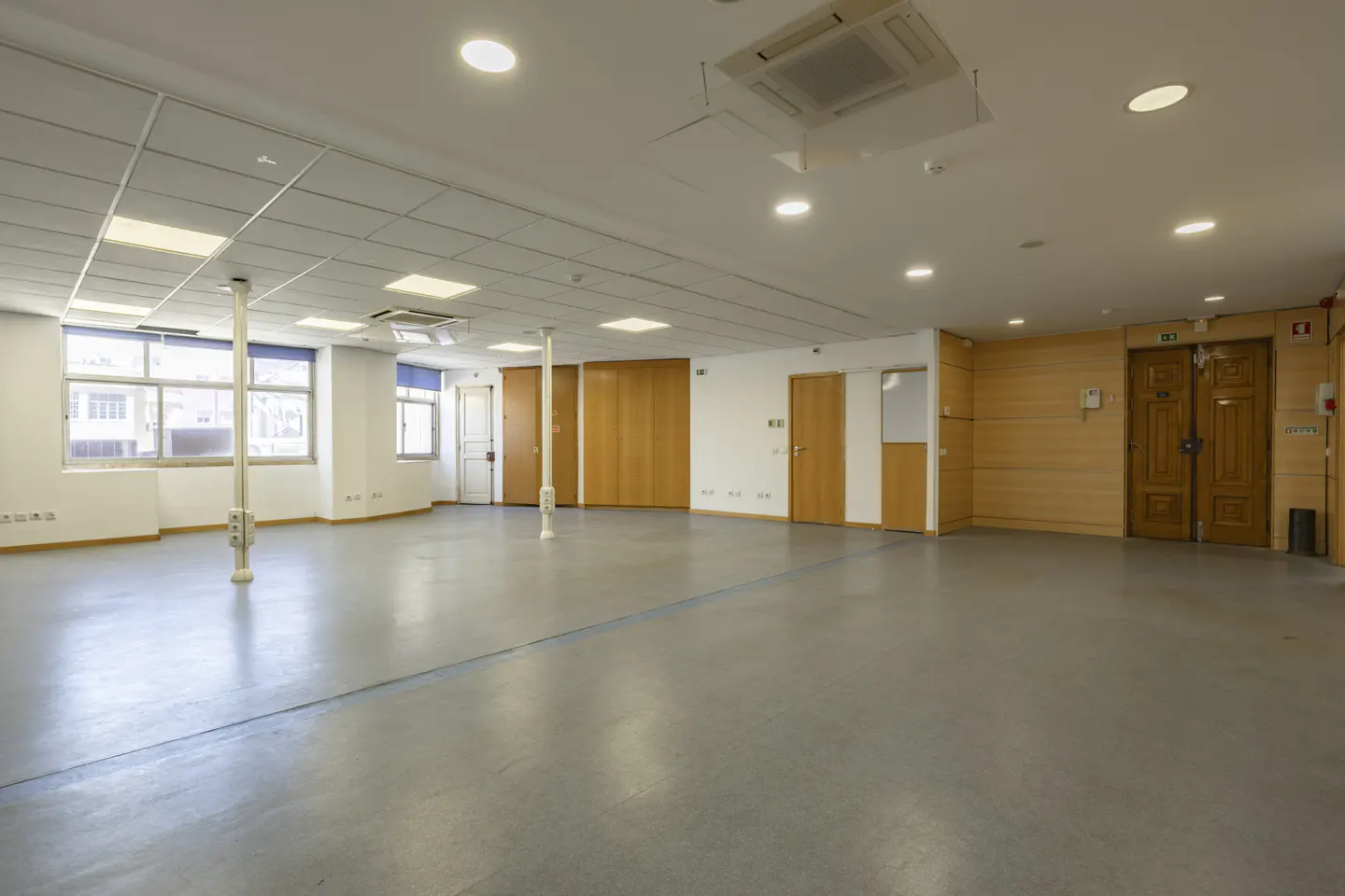 A large, empty room with gray floors, white walls, and a white ceiling with recessed lighting. There are wooden doors and support columns throughout the space.
