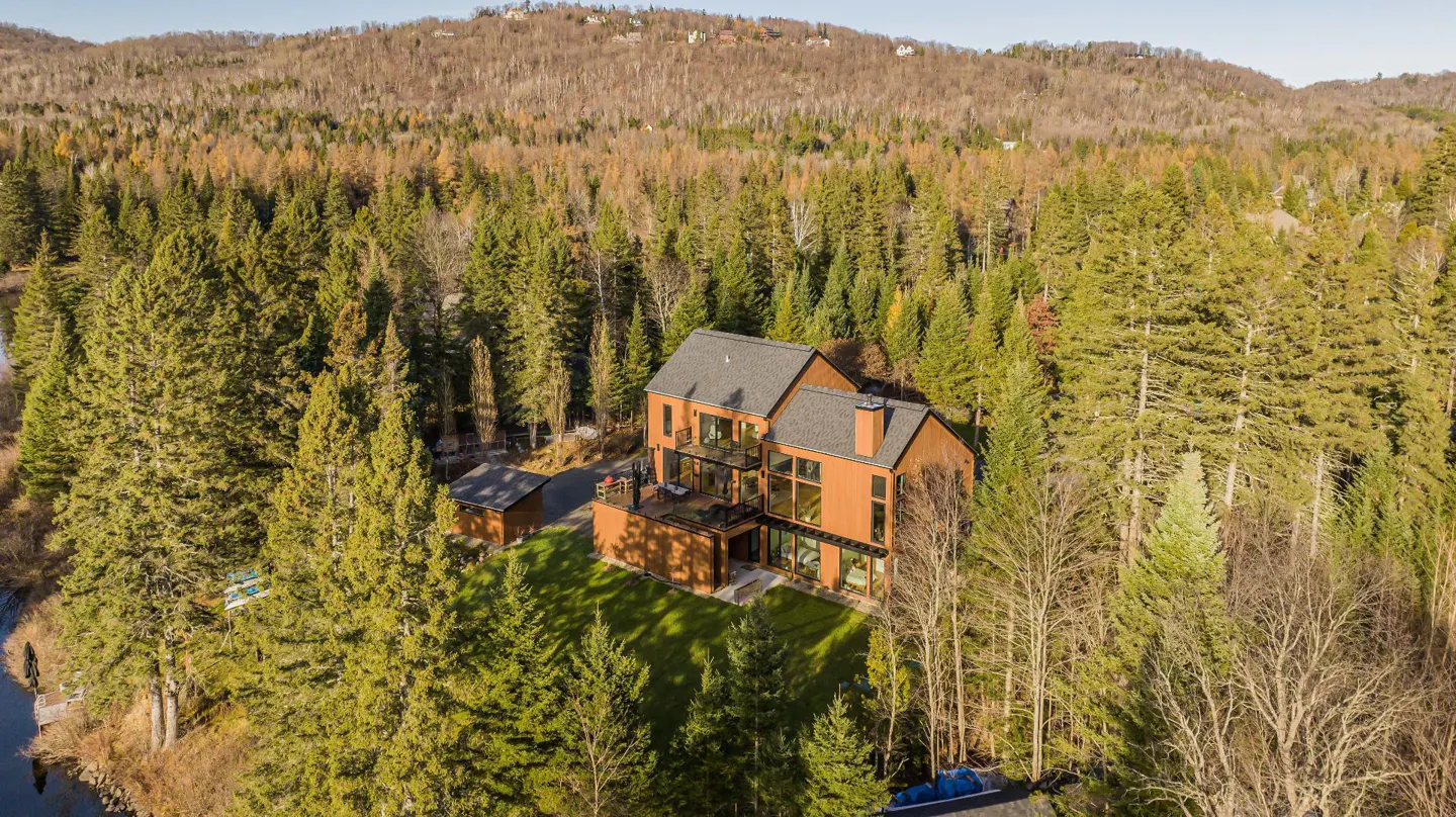 Aerial view of a modern brown house with a gray roof, surrounded by green trees and a mountain in the background.