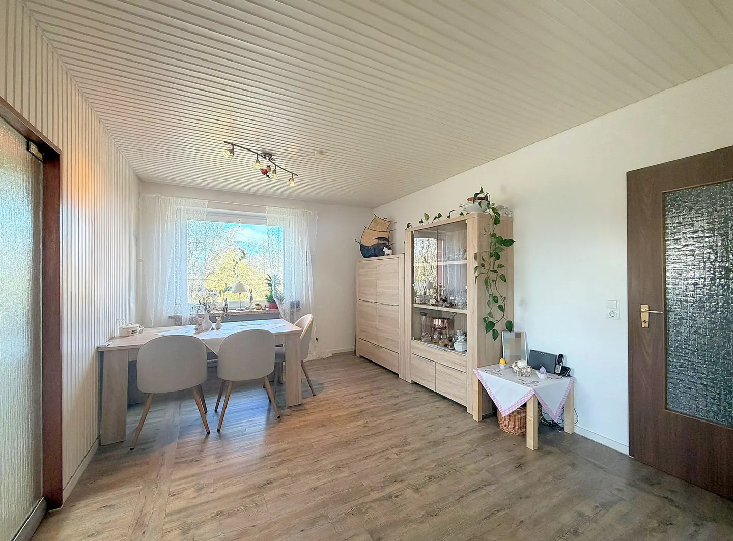A bright dining room with a table, chairs, and a display cabinet. A window with sheer curtains lets in natural light. The floor is light wood-toned.