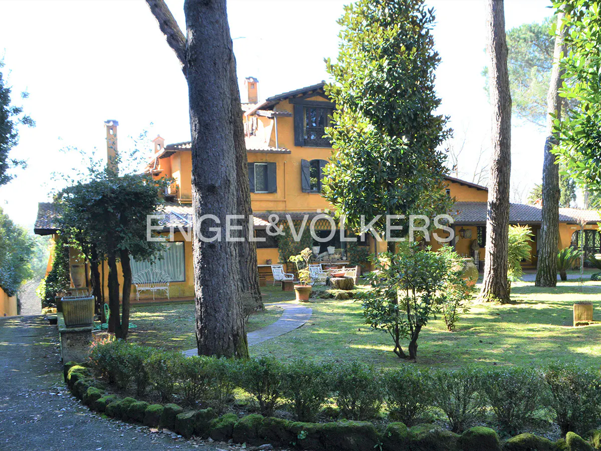 A yellow villa with dark shutters sits in a lush garden with tall pine trees, a stone path, and a manicured hedge.