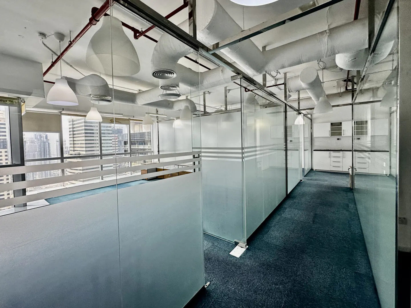 Office space with glass-walled cubicles and blue carpet. White ceiling with exposed ductwork and modern lighting. Cityscape visible through windows.