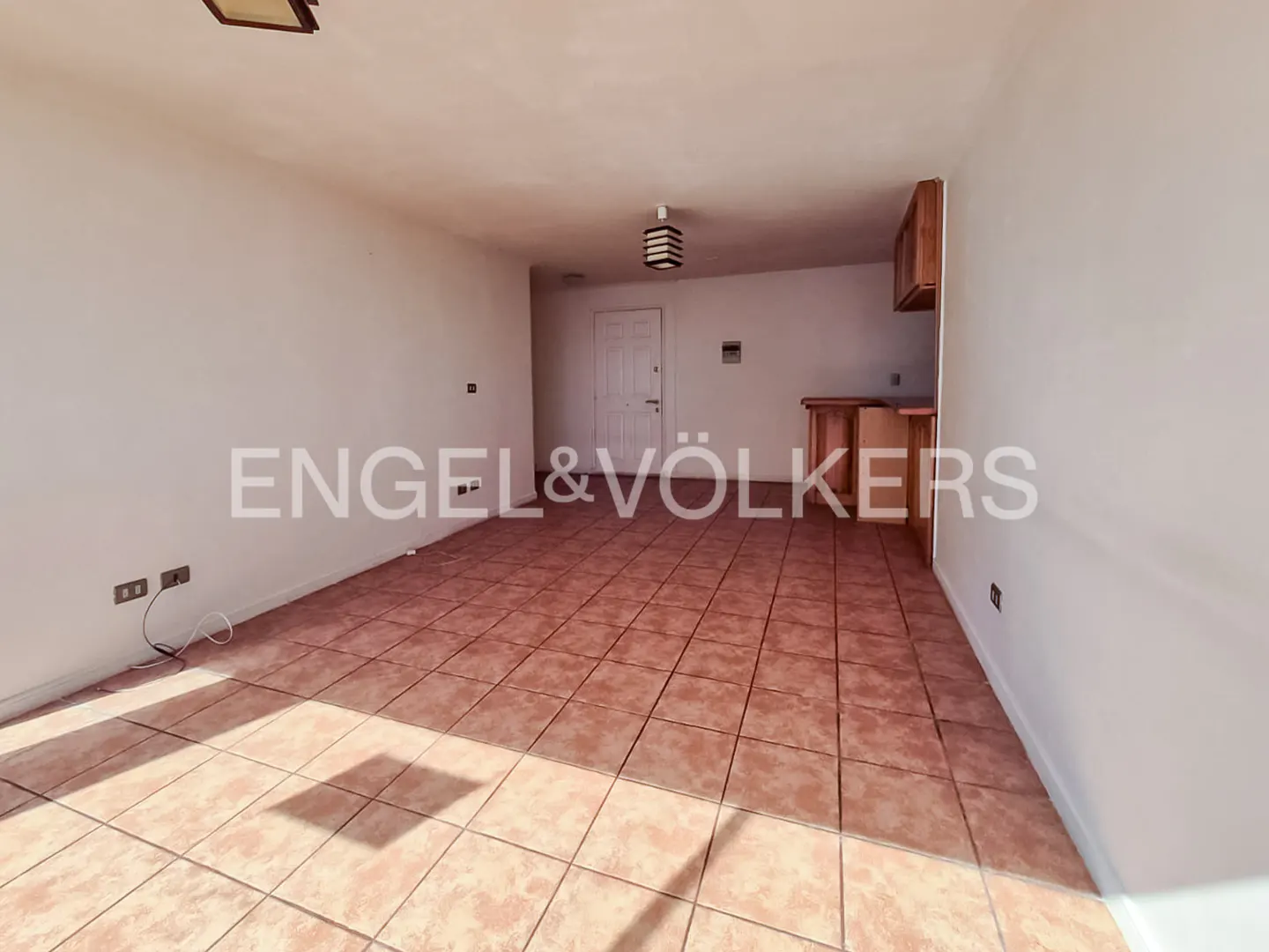 An empty room with white walls, brown tile floor, and a small kitchen area in the back.