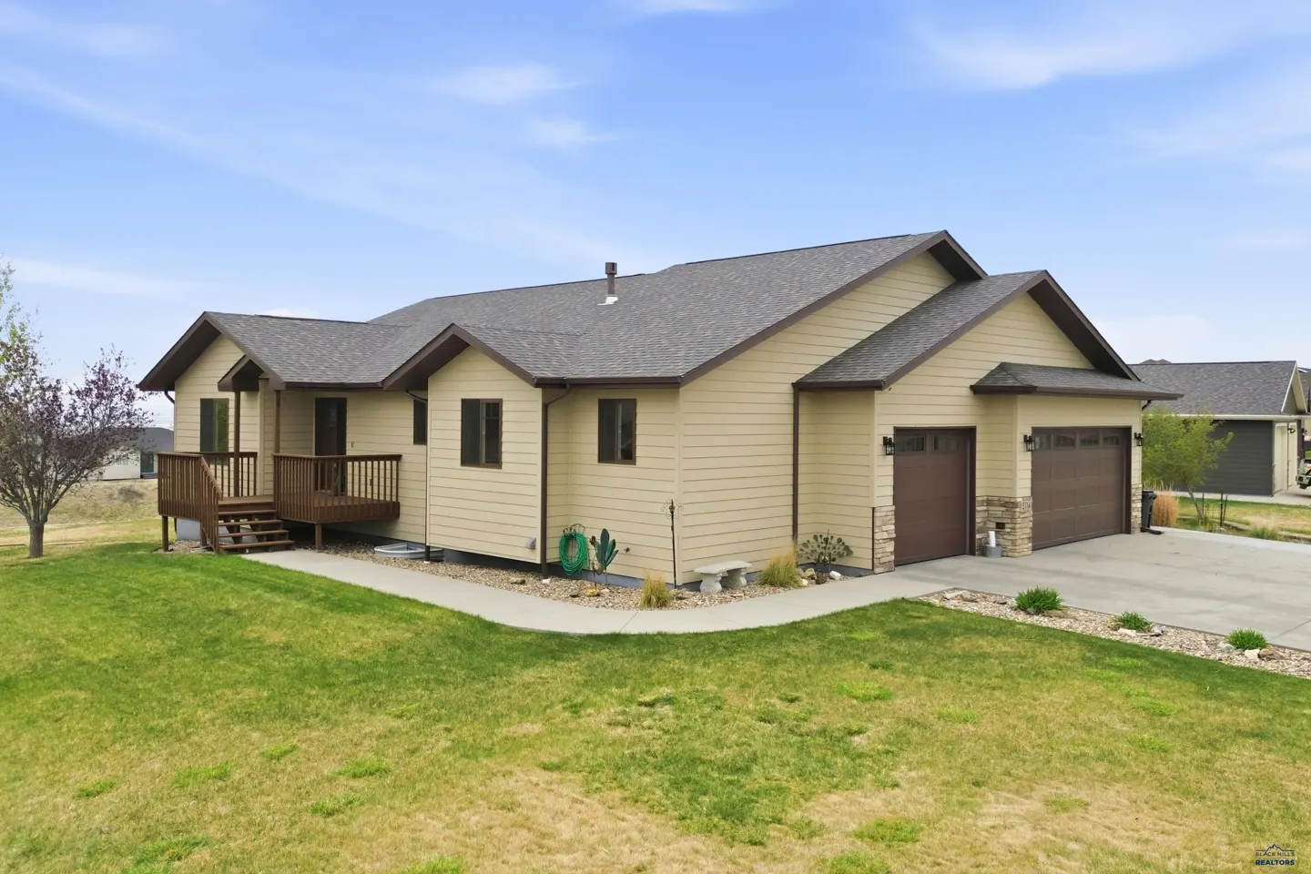 Tan single-story house with a brown roof, brown trim, and a two-car garage on a green lawn under a blue sky.