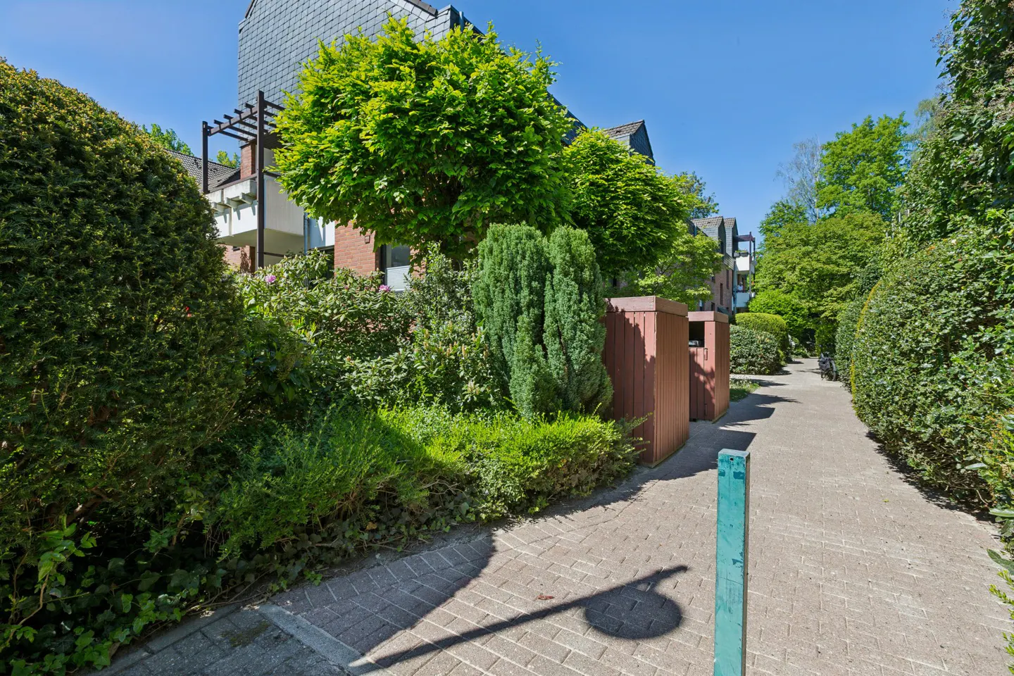 Brick walkway lined with lush green bushes and trees leading to apartments on a sunny day.