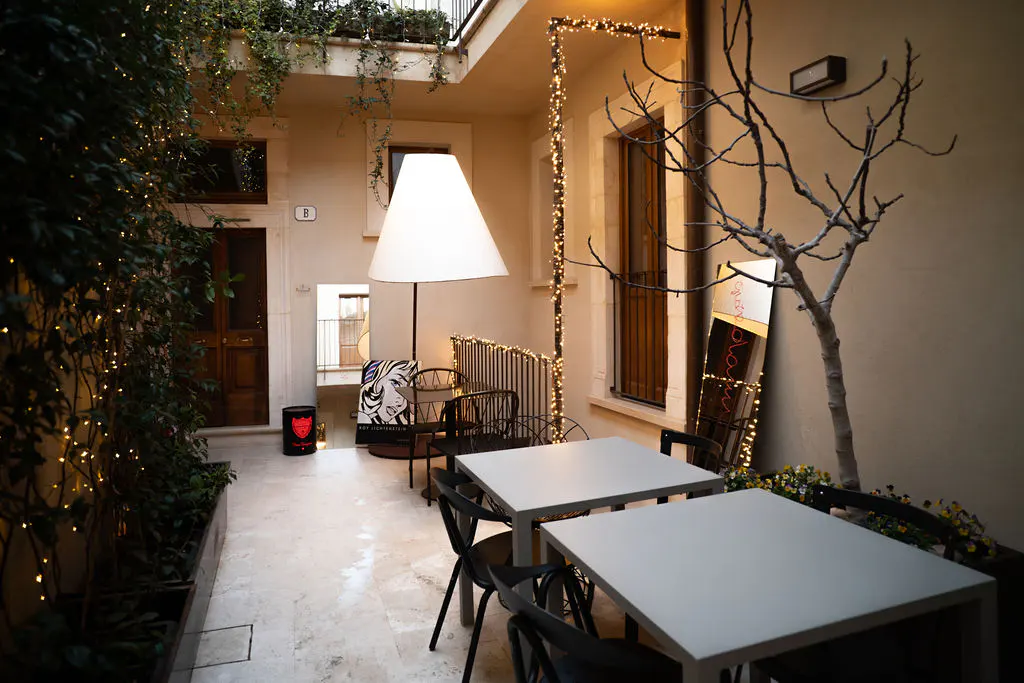 Outdoor patio with white tables, black chairs, and string lights. A large white lamp and artwork add to the ambiance.