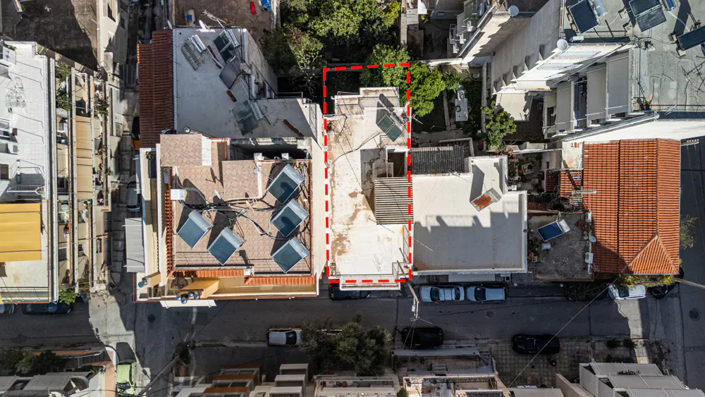 Aerial view of a property outlined in red, surrounded by buildings, trees, and parked cars. Solar panels are visible on rooftops.