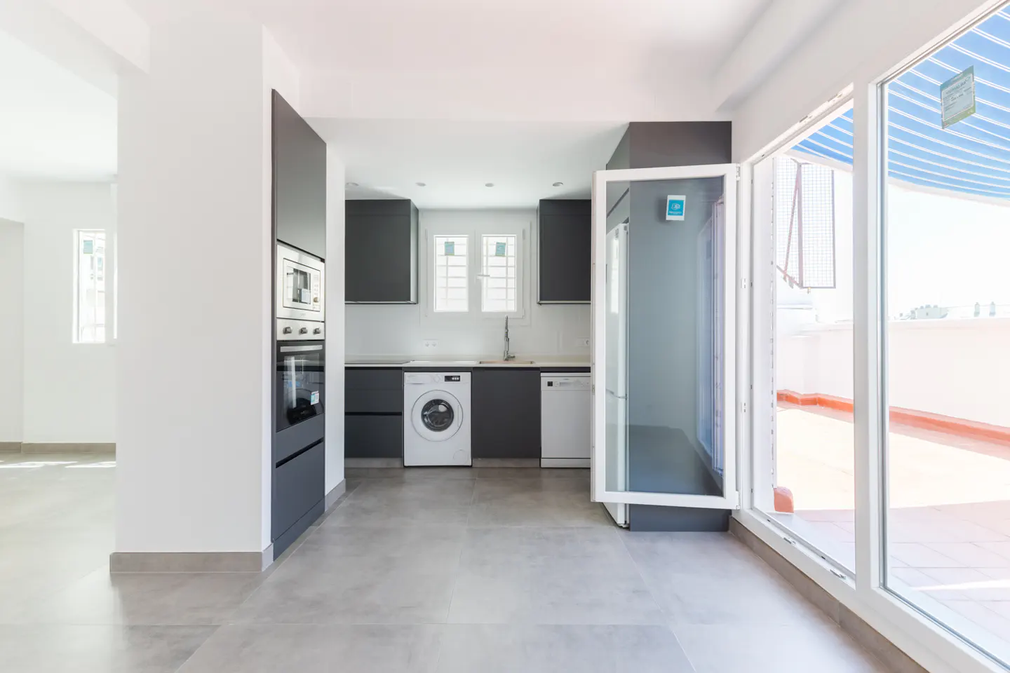 Modern kitchen with dark grey cabinets, built-in appliances, and a washing machine, opening onto a bright outdoor terrace.