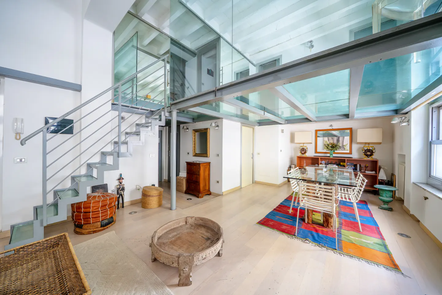 Bright, modern living room with glass-floored mezzanine. Metal stairs lead up. Dining table with colorful rug. Light wood floors.