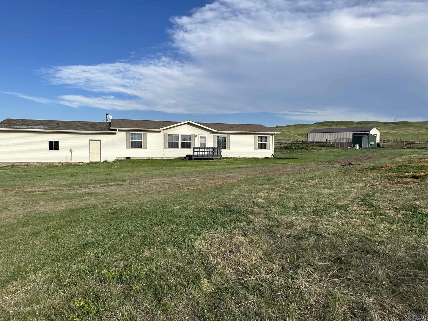 Single-story beige home with a brown roof, small deck, and detached garage on a large grassy lot under a blue sky.