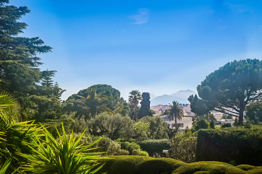 Lush green garden with palm trees and manicured hedges under a clear blue sky, overlooking a distant cityscape and mountains.