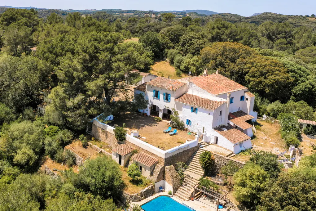 Aerial view of a white villa with a terracotta roof, blue shutters, a courtyard, and a swimming pool in a wooded area.