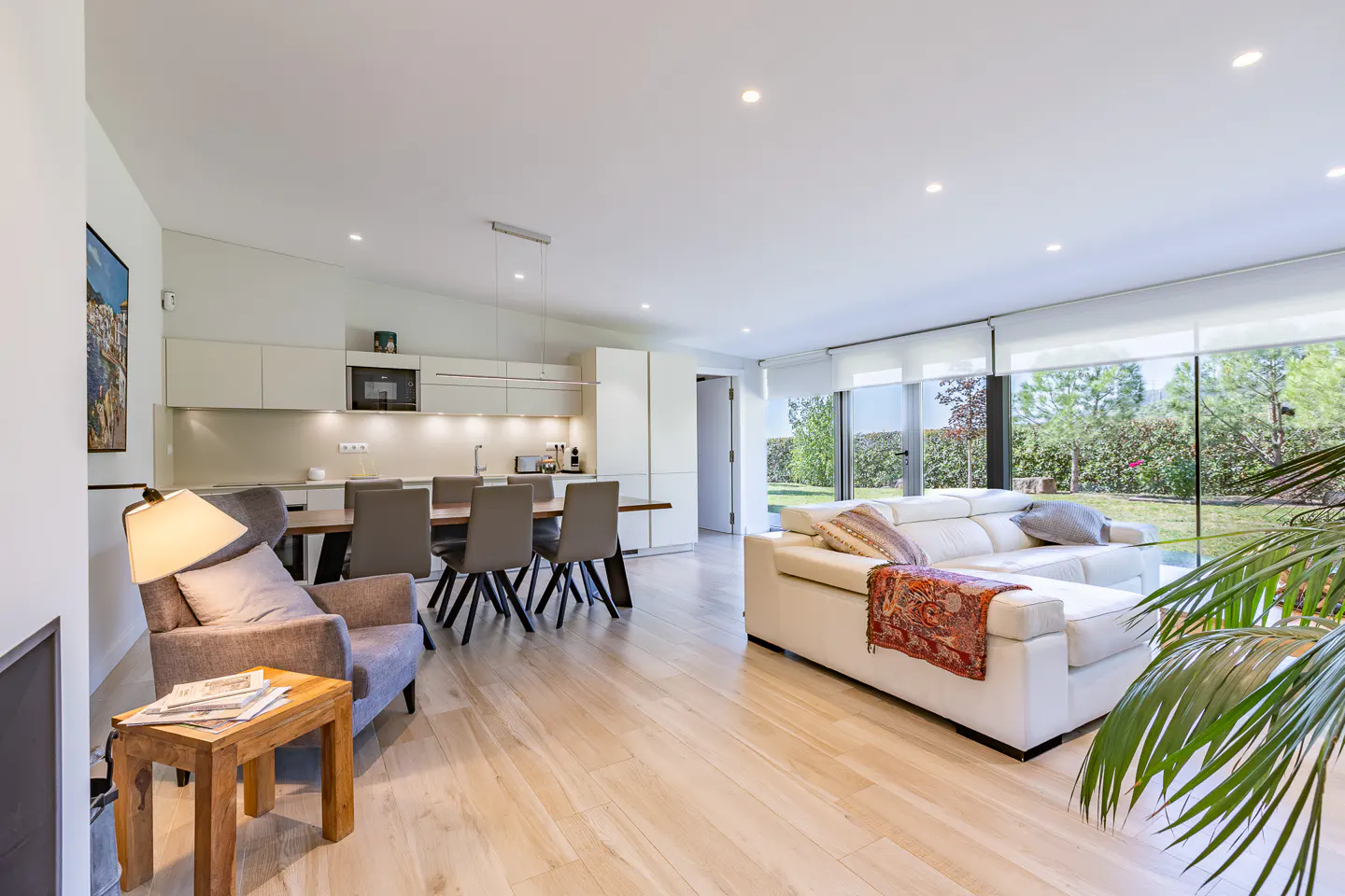 Bright, open-plan living space with light wood floors, white walls, and a modern kitchen. A white sofa faces a large window overlooking a green garden.