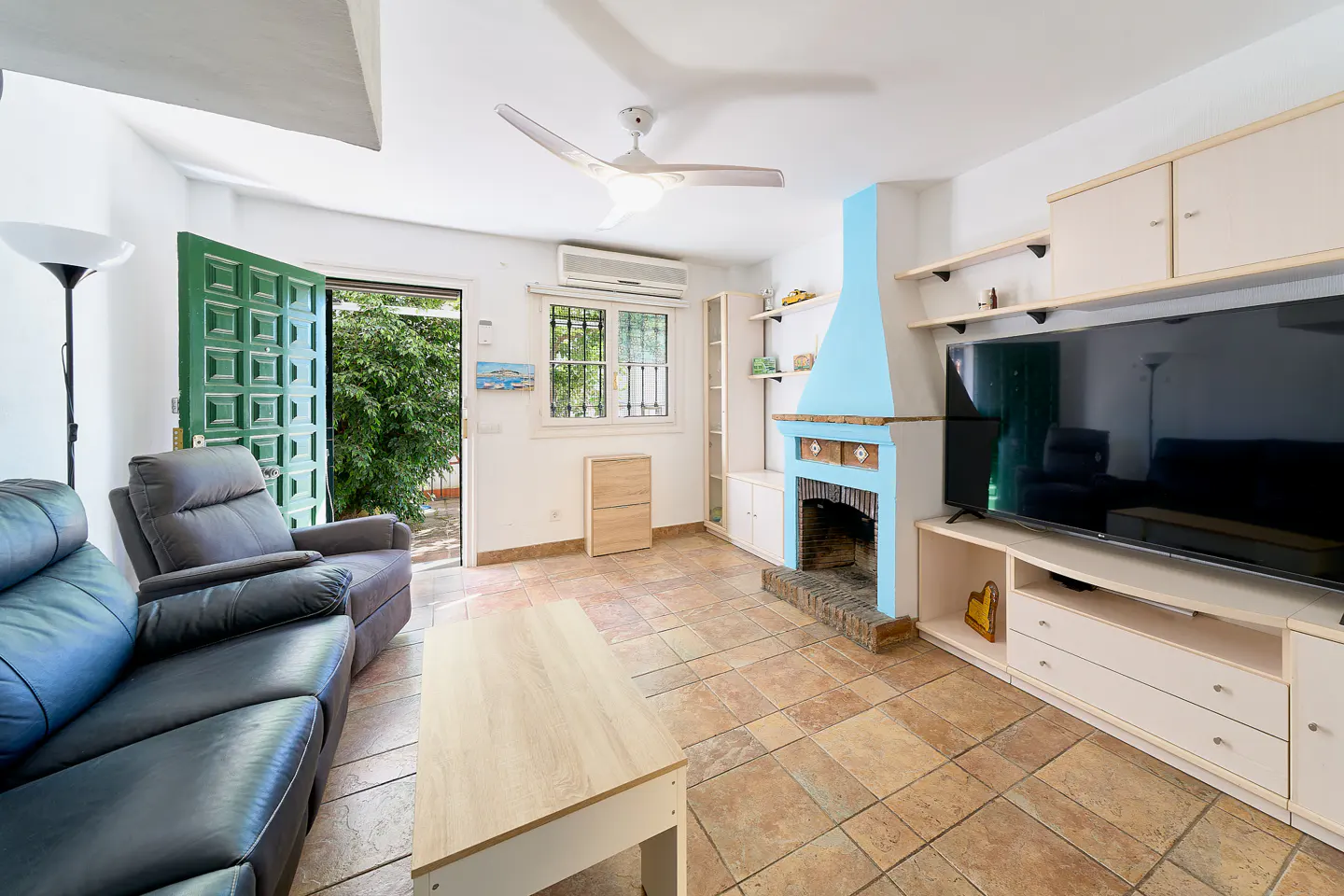 Bright living room with a blue fireplace, a large TV, and a green door leading to a garden. A gray sofa and a light wood coffee table are in the foreground.
