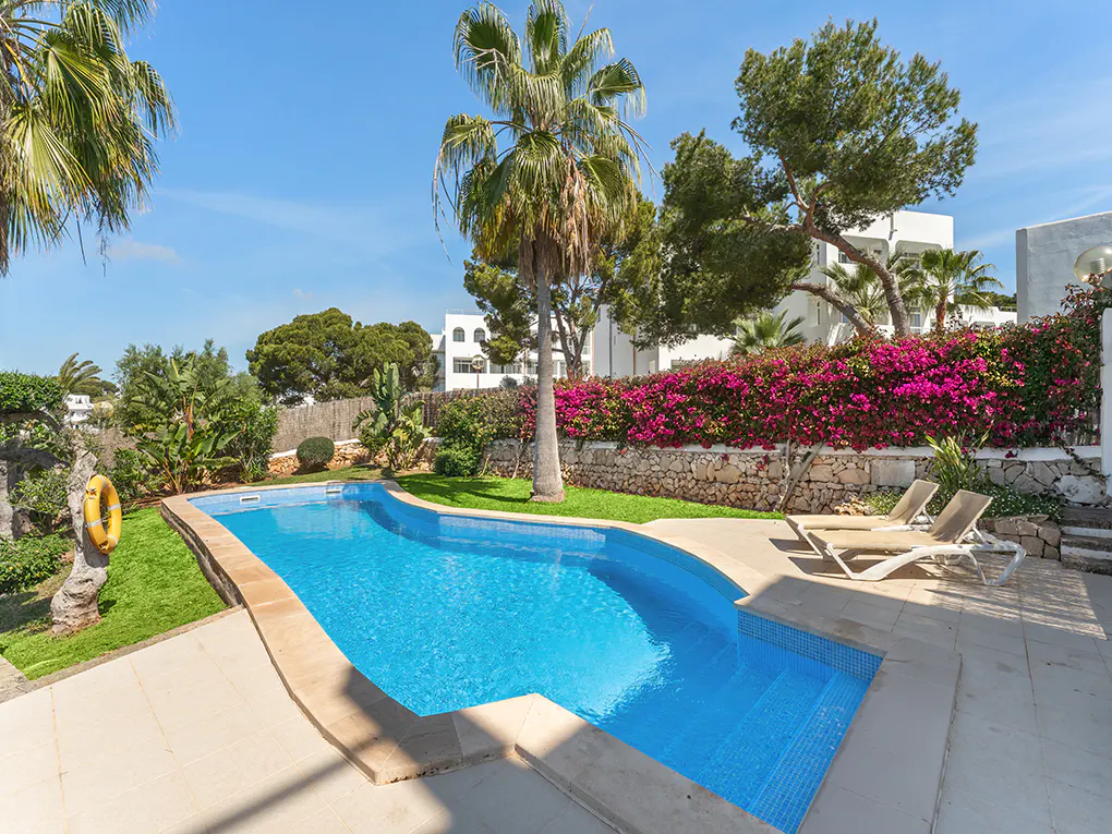 A bright blue swimming pool is surrounded by lush greenery, palm trees, and a white building under a clear blue sky. Two lounge chairs sit on the patio.