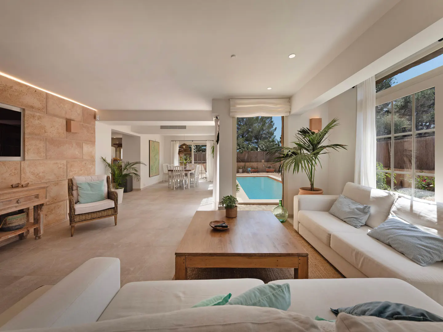 Bright, open living room with white sofas, wood table, and pool view. Stone wall and dining area visible.