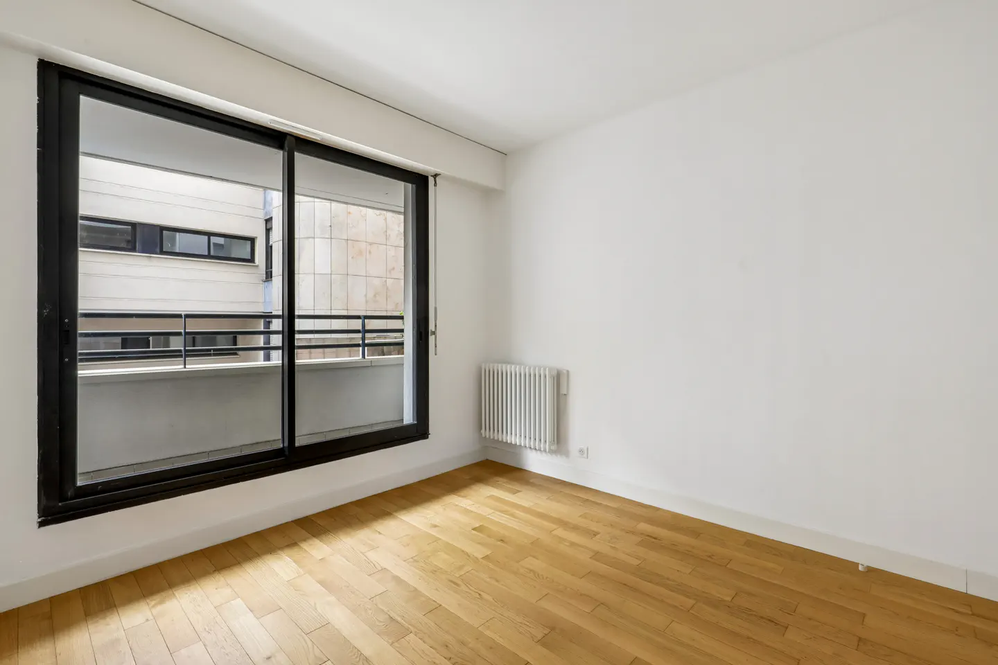 Bright, empty room with wood floors, white walls, and a black-framed sliding glass door to a balcony. A white radiator is on the wall.