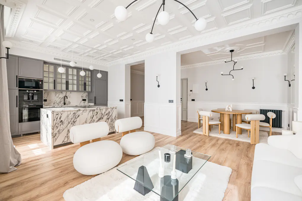 Open-concept living space with a marble island, gray cabinets, and white seating. A dining area is visible in the background.